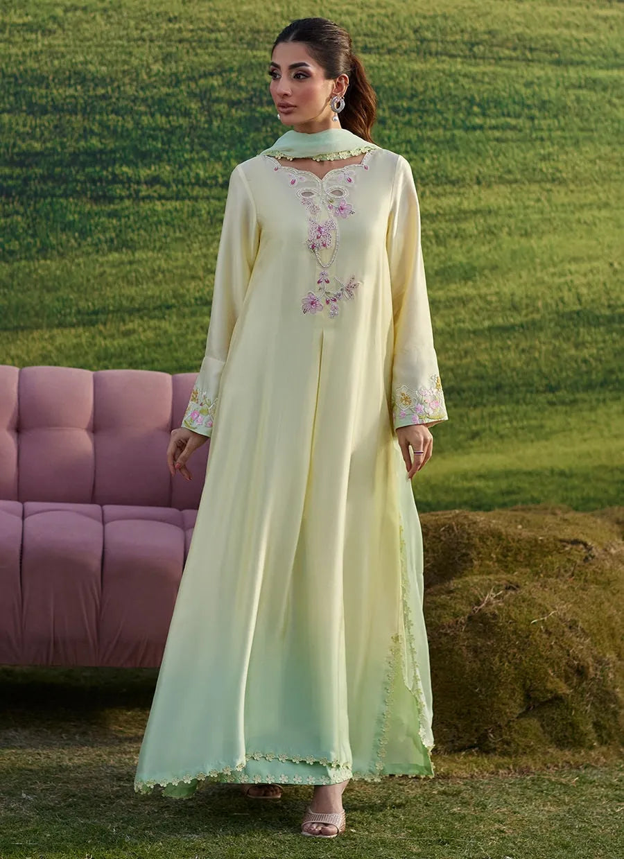 Woman in a light green embroidered dress standing outdoors with a pink sofa and grassy background.