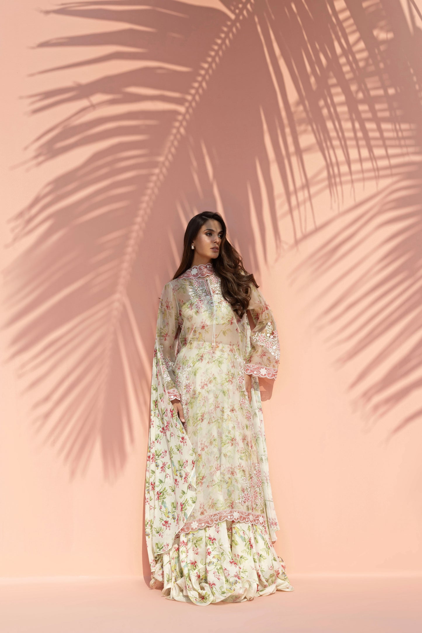 Woman in a floral dress standing against a pink background with palm leaf shadows