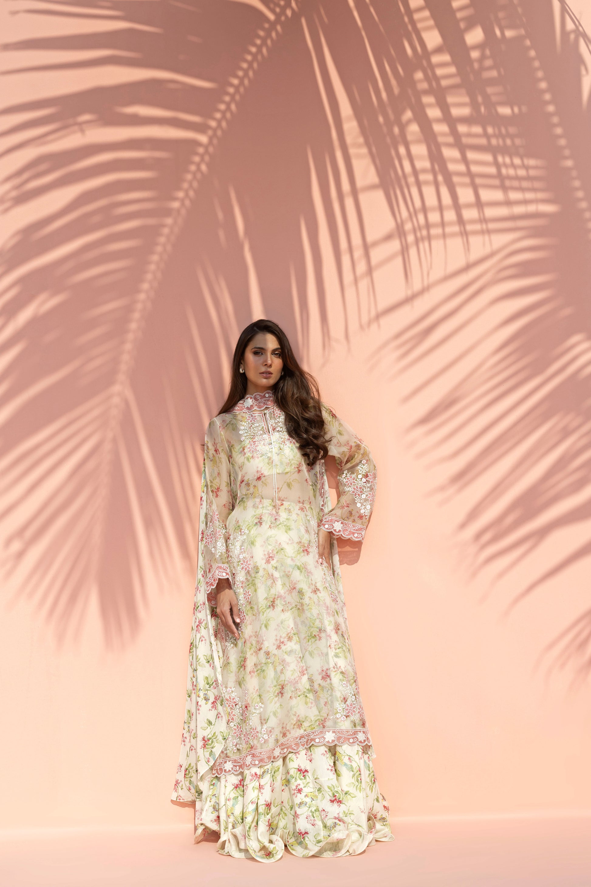 Woman in a floral dress standing against a pink wall with palm leaf shadows