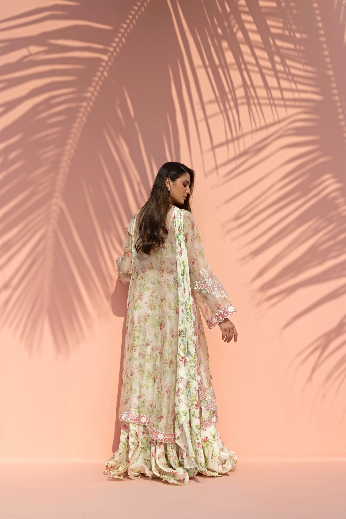 Woman in a floral dress standing against a pink background with palm leaf shadows