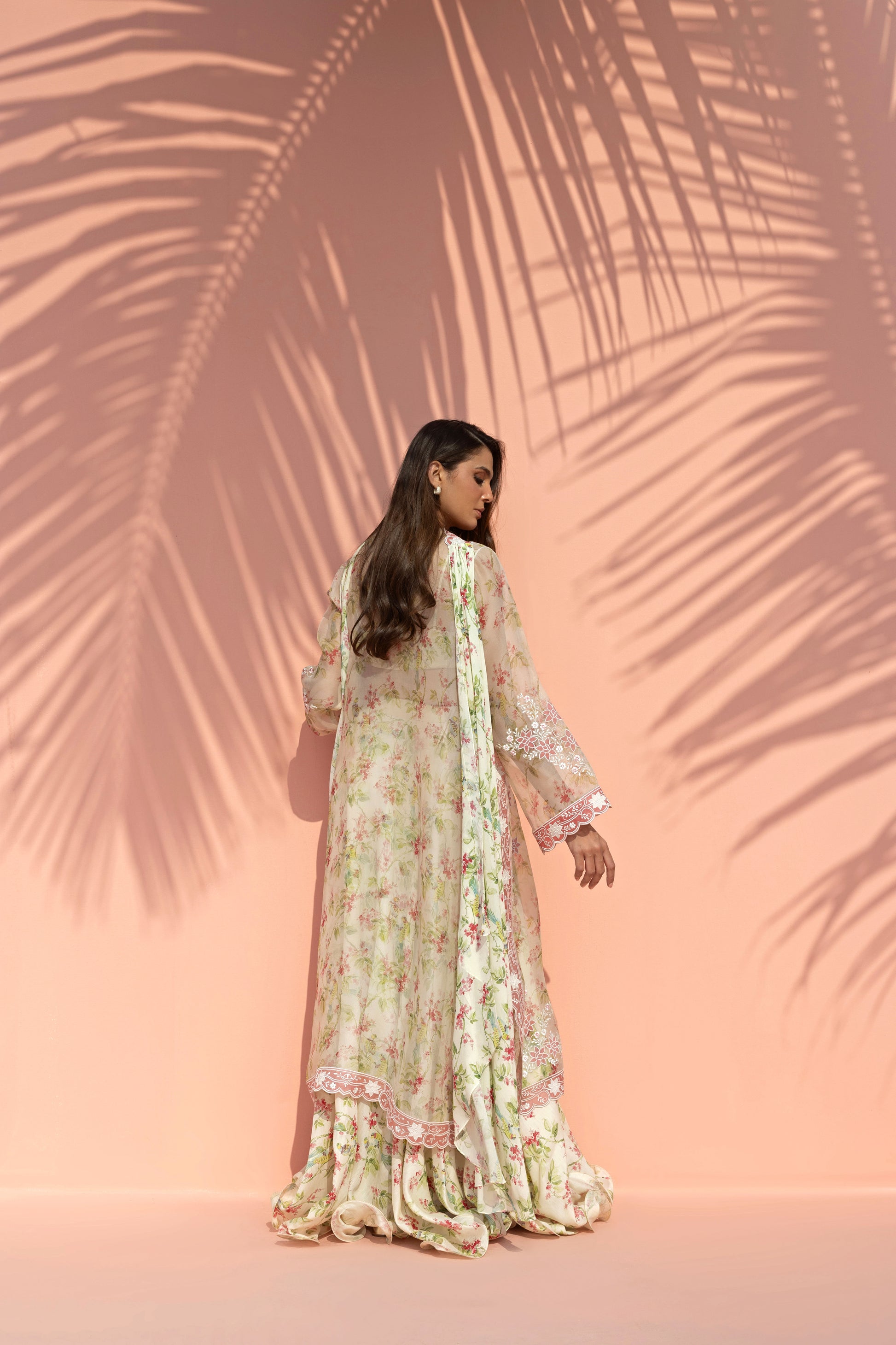 Woman in a floral dress standing against a pink background with palm leaf shadows