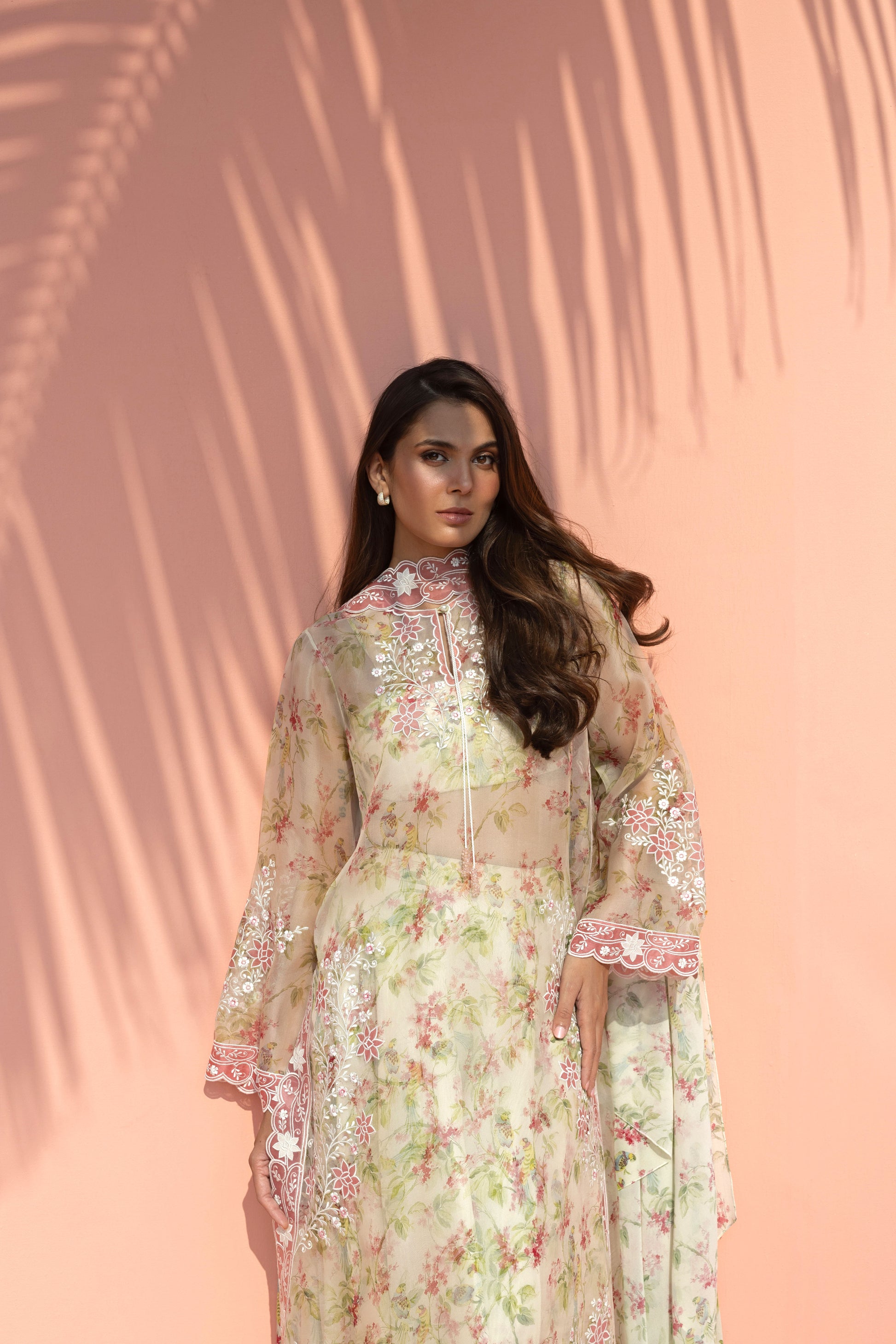 Woman in a floral dress standing against a pink background with palm leaf shadows.