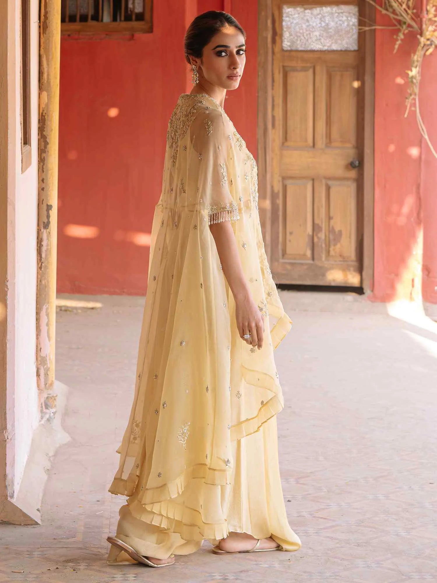 Woman in a yellow traditional outfit standing in a sunlit room with wooden door and red wall.