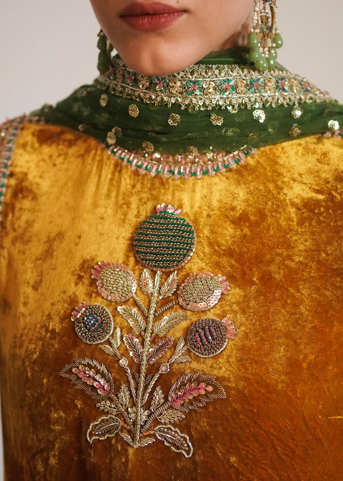 Close-up of a person wearing a mustard yellow outfit with floral embroidery and green jewelry.