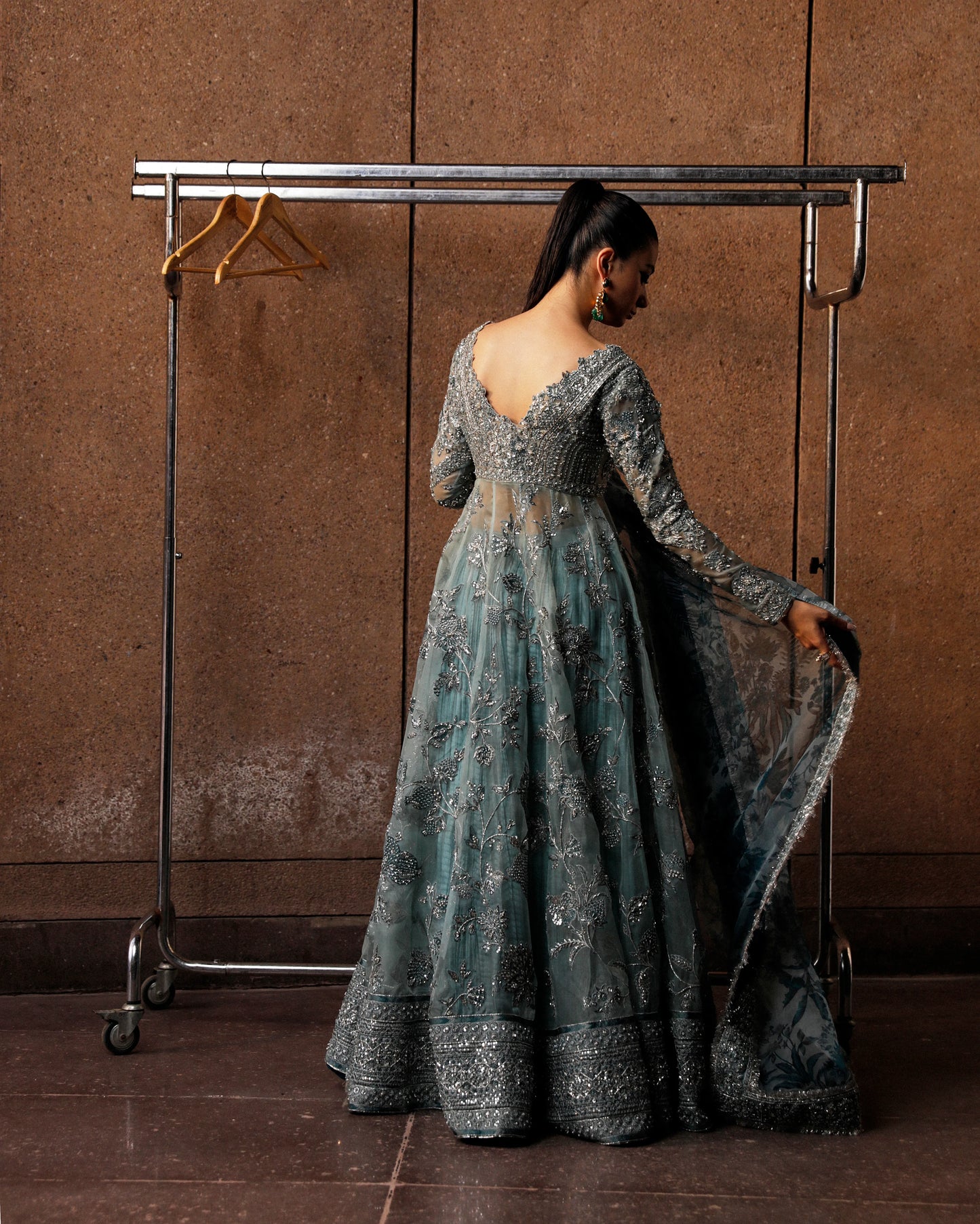 Woman in a long, elegant dress standing next to a clothing rack against a brown wall.