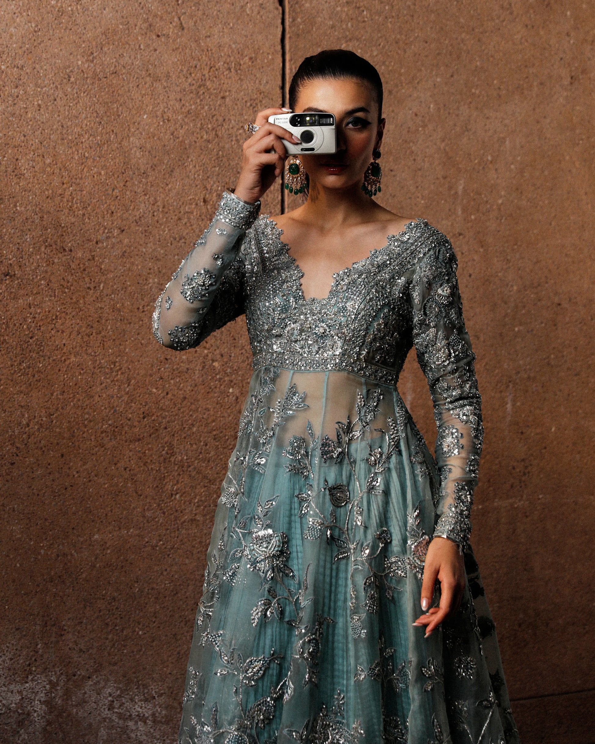 Woman in a silver and blue embroidered dress taking a mirror selfie against a brown background