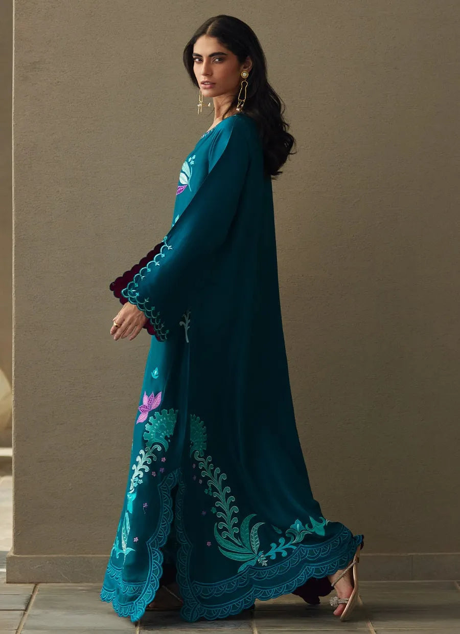 Woman wearing a teal embroidered dress against a beige wall.