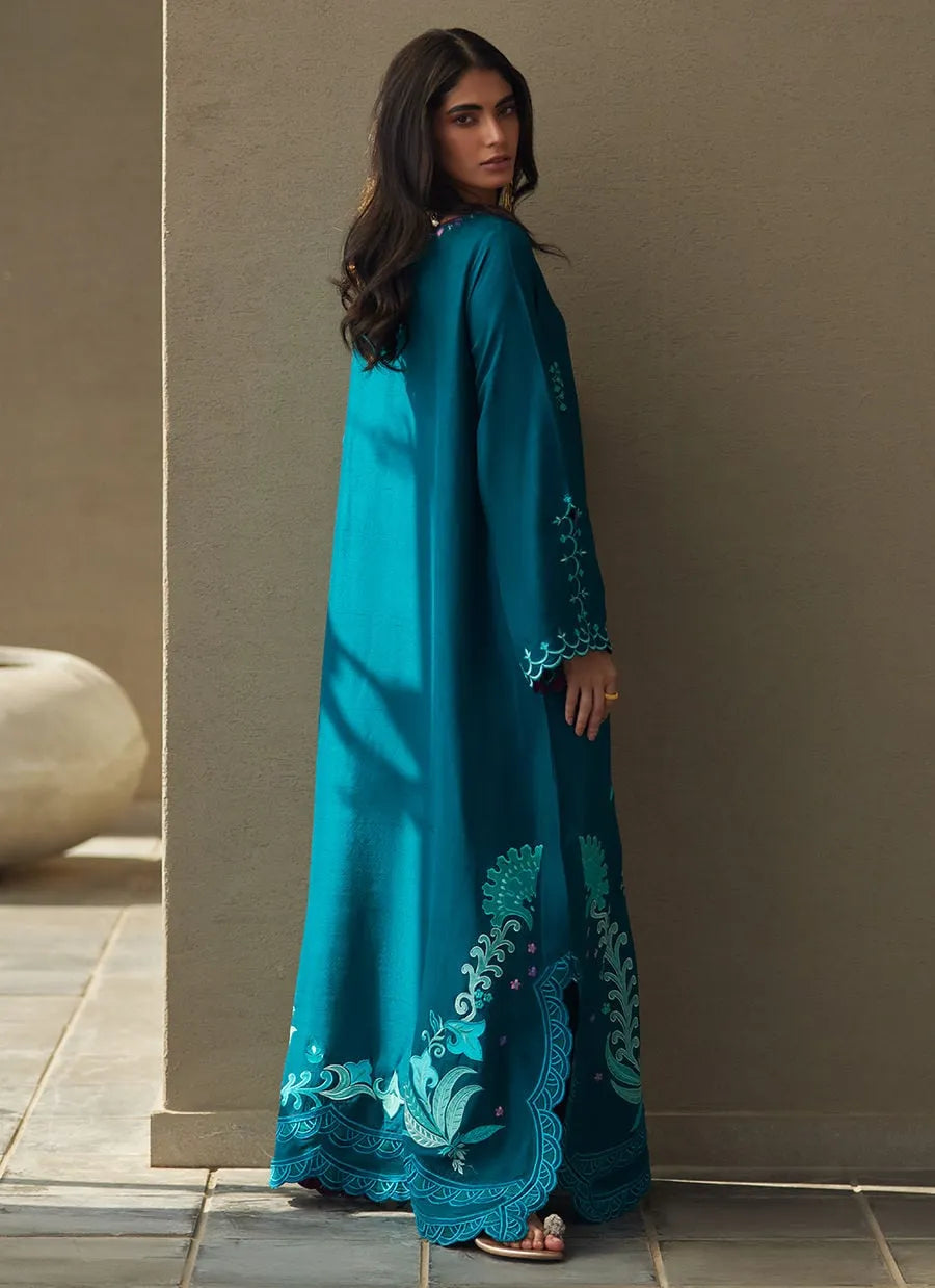 Woman wearing a turquoise embroidered dress against a beige wall.