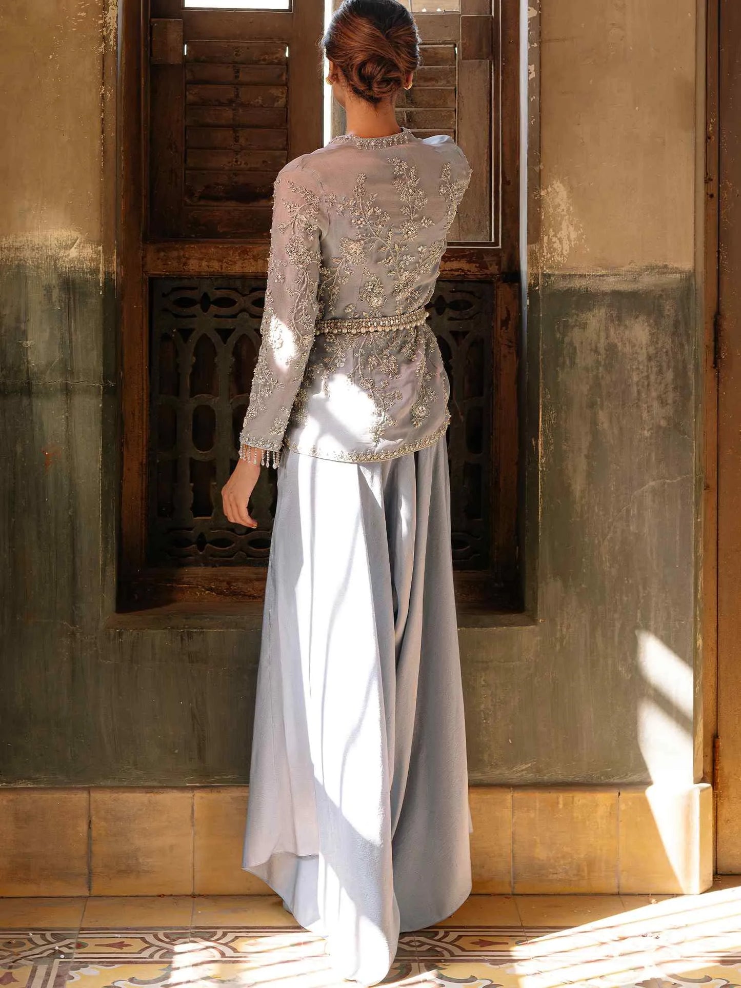 Woman wearing a light gray traditional outfit standing in a sunlit room with wooden walls.