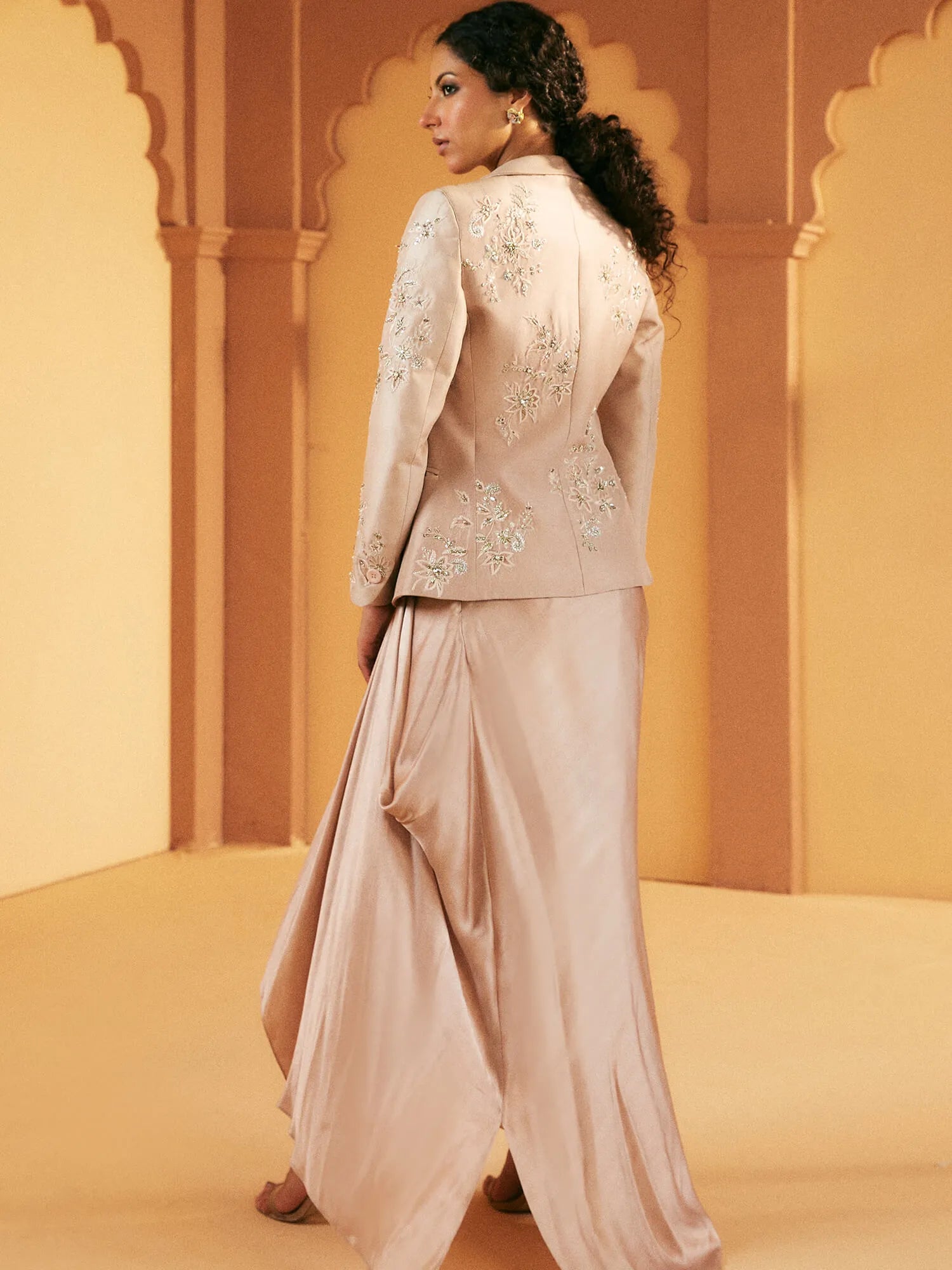 Woman wearing a traditional outfit with an embroidered jacket and flowing pants in a warm-toned setting.