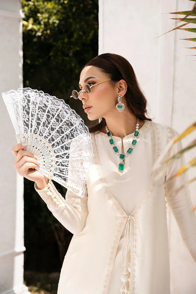 Woman in a white outfit holding a lace fan, wearing green jewelry, with a blurred outdoor background.