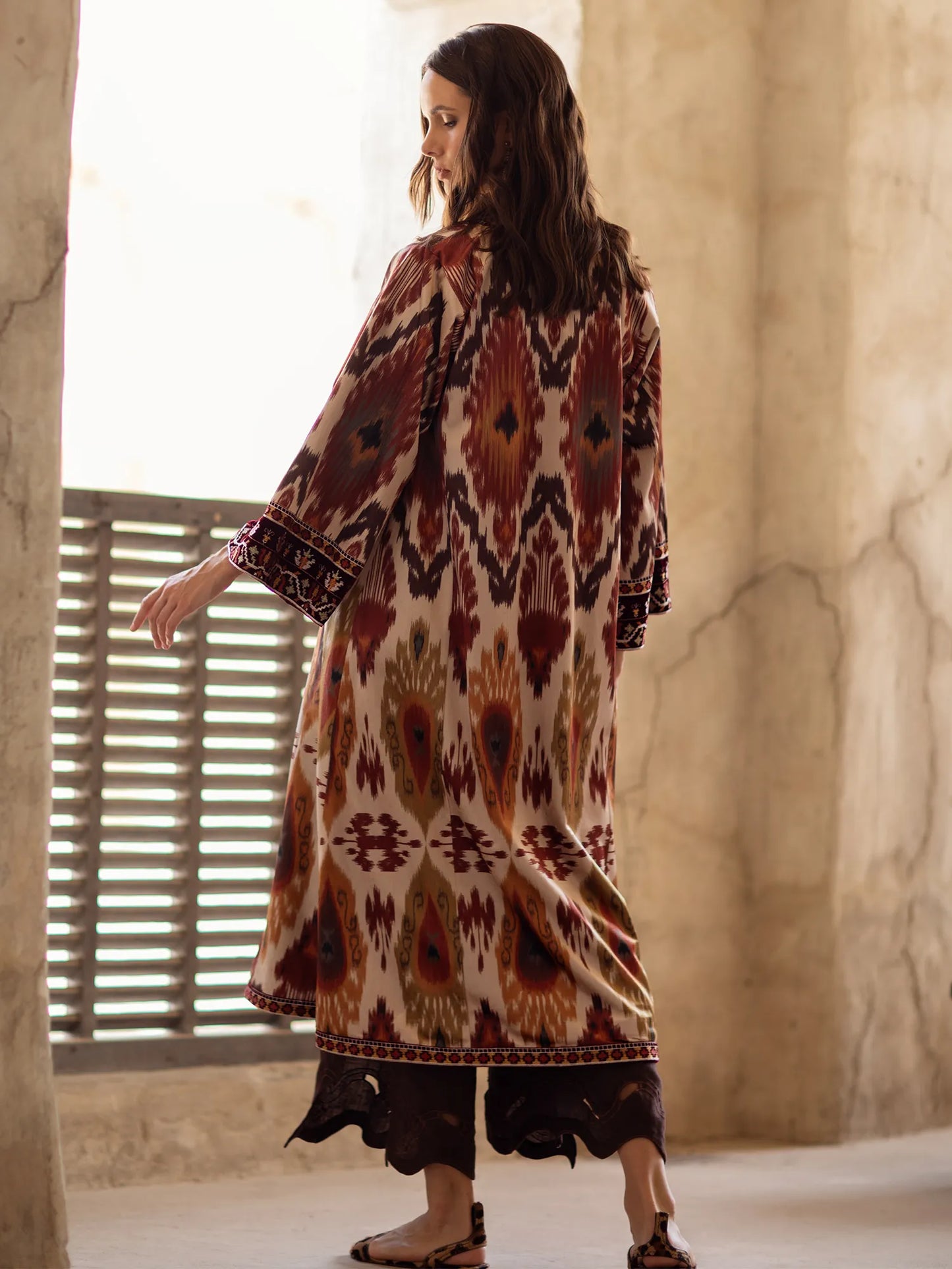 Woman wearing a patterned dress standing in a sunlit room with stone walls.
