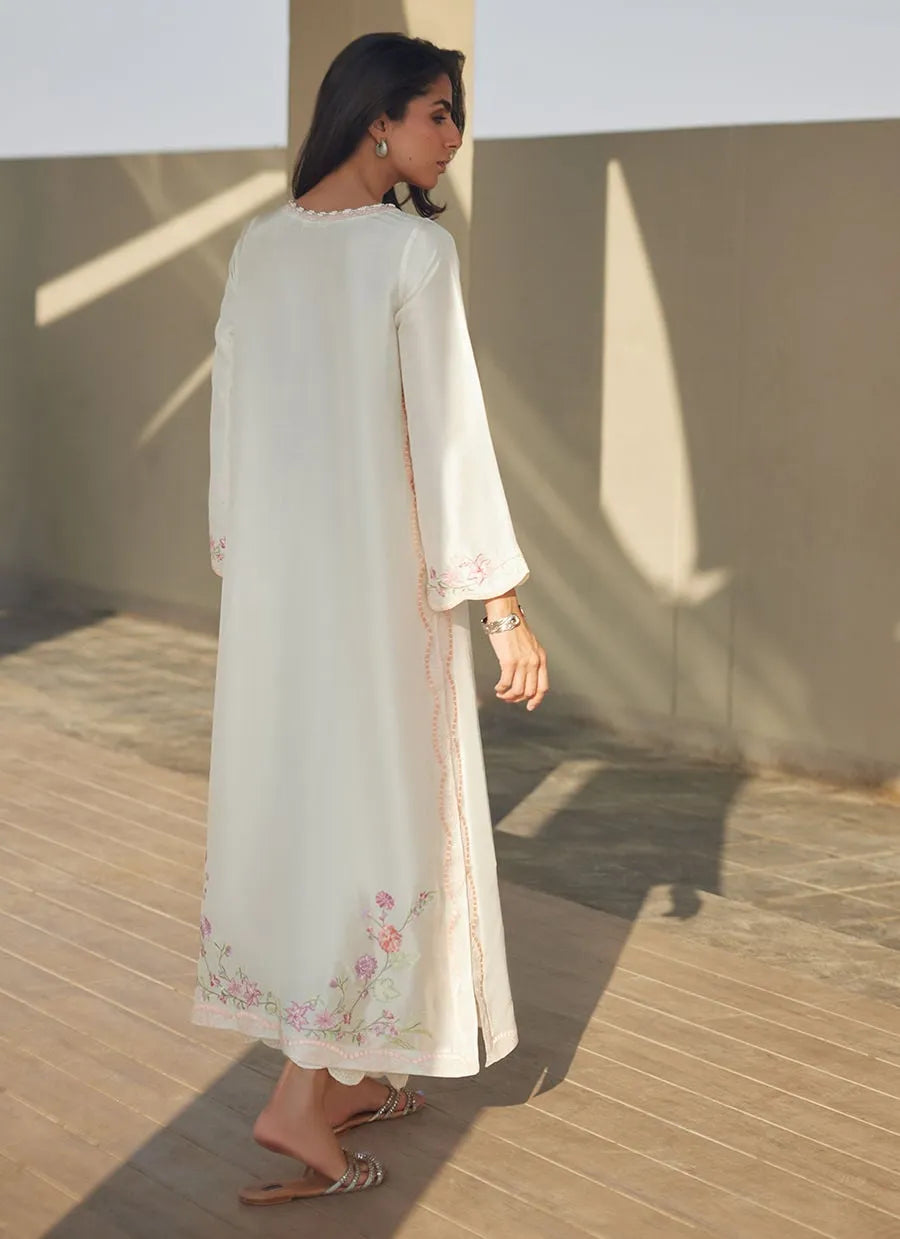 Woman wearing a long, light-colored dress with floral embroidery on a wooden deck.