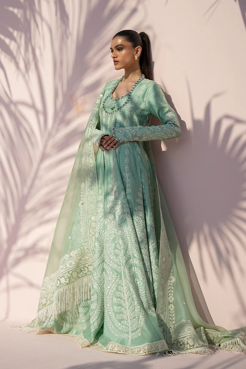 Woman wearing a light green traditional outfit with intricate patterns against a white wall.