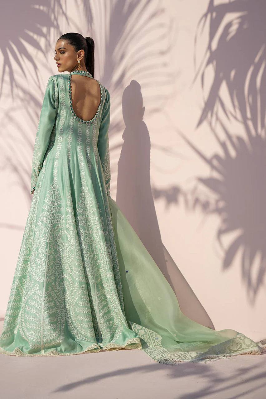 Woman wearing a light green traditional outfit with a backless design, standing against a neutral background with palm leaf shadows.