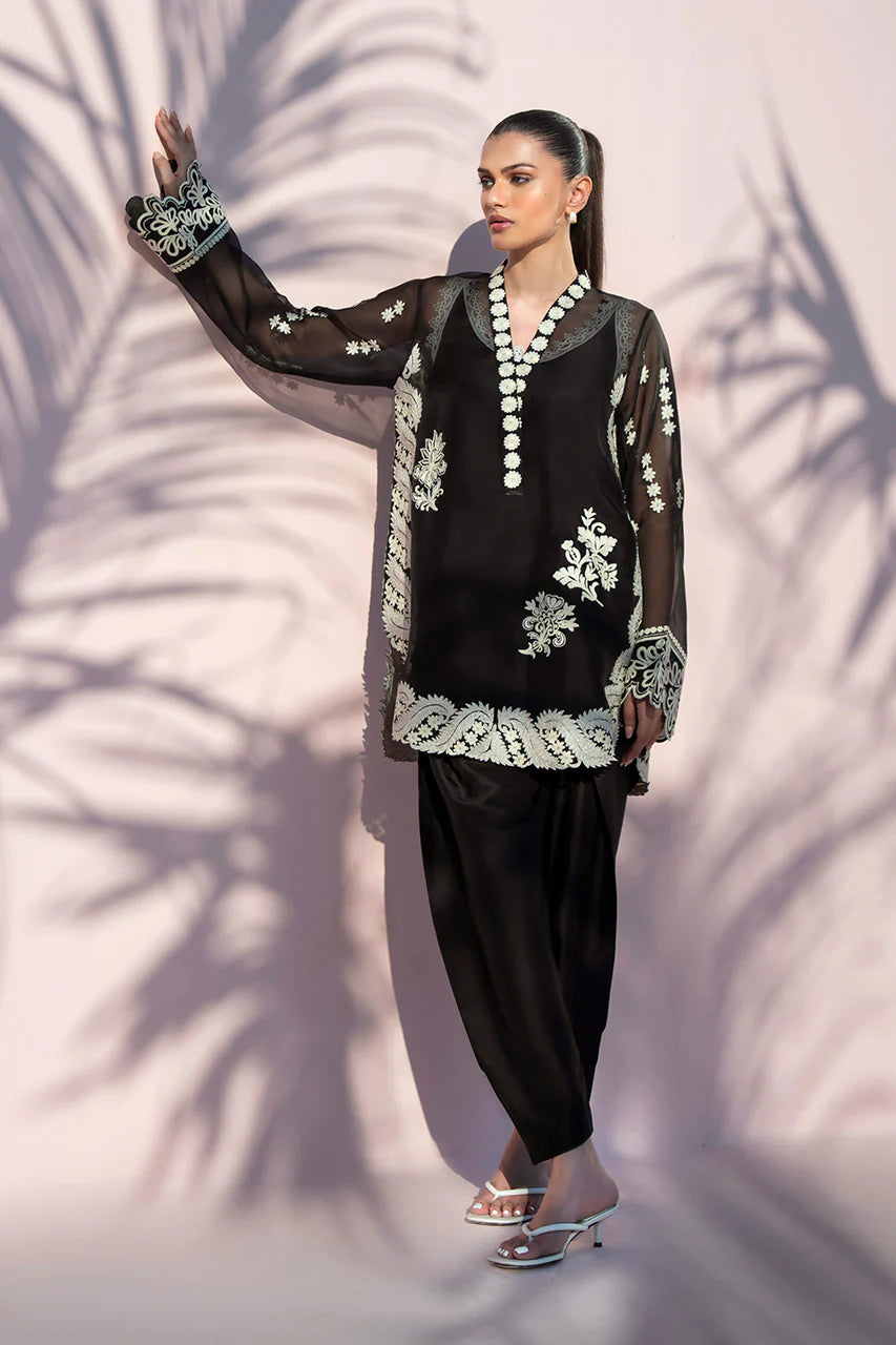 Woman in a black and white embroidered outfit standing against a textured wall.