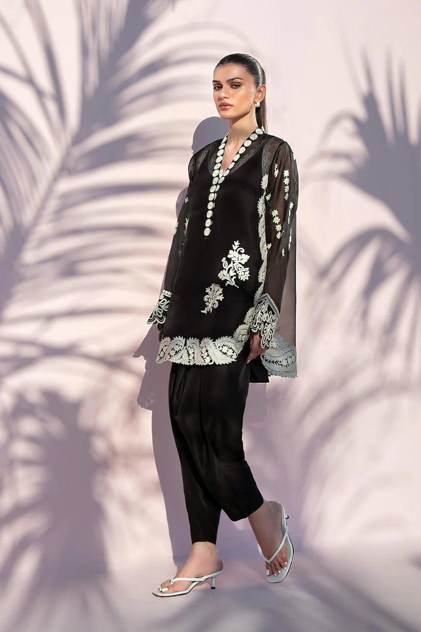 Woman wearing a black outfit with white embroidery against a textured wall.