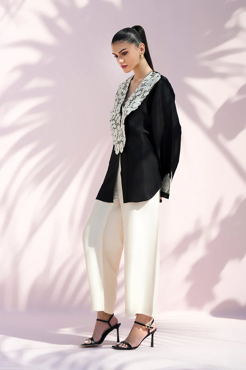 Woman wearing a black blazer with floral details, white pants, and high heels against a light pink background.