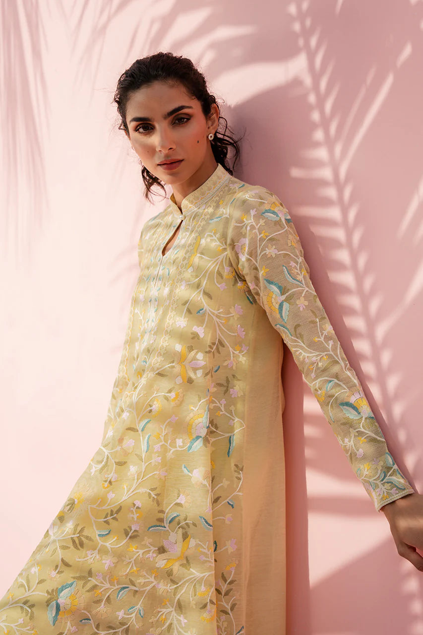 Woman wearing a light yellow floral dress against a pink wall with palm leaf shadows.
