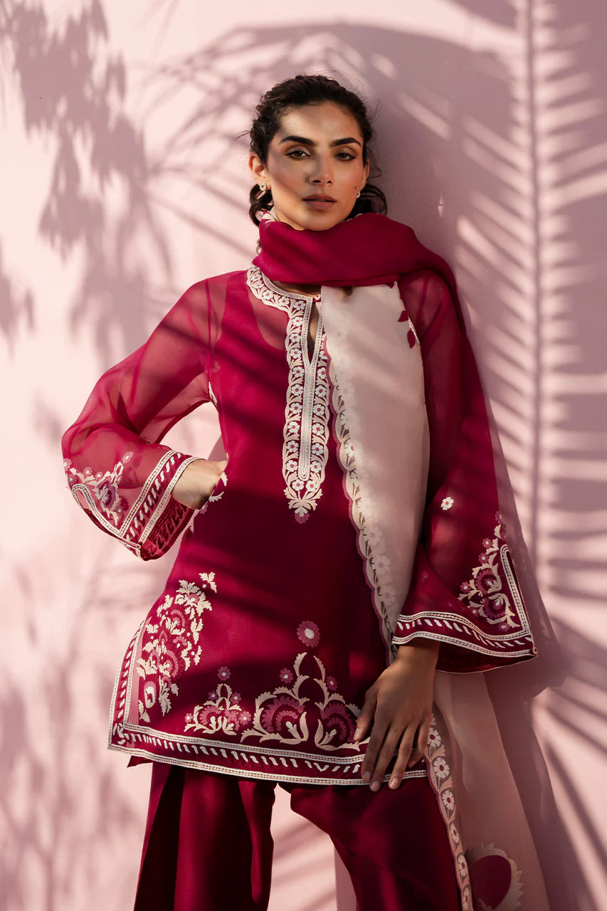 Woman wearing a red traditional outfit with white embroidery against a pink background