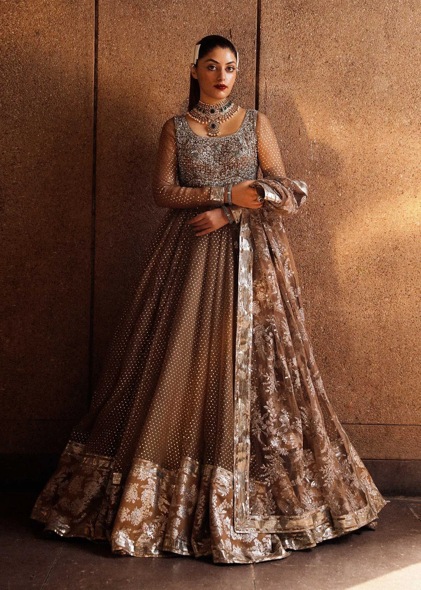 Woman in an elegant traditional outfit with intricate designs against a textured wall.