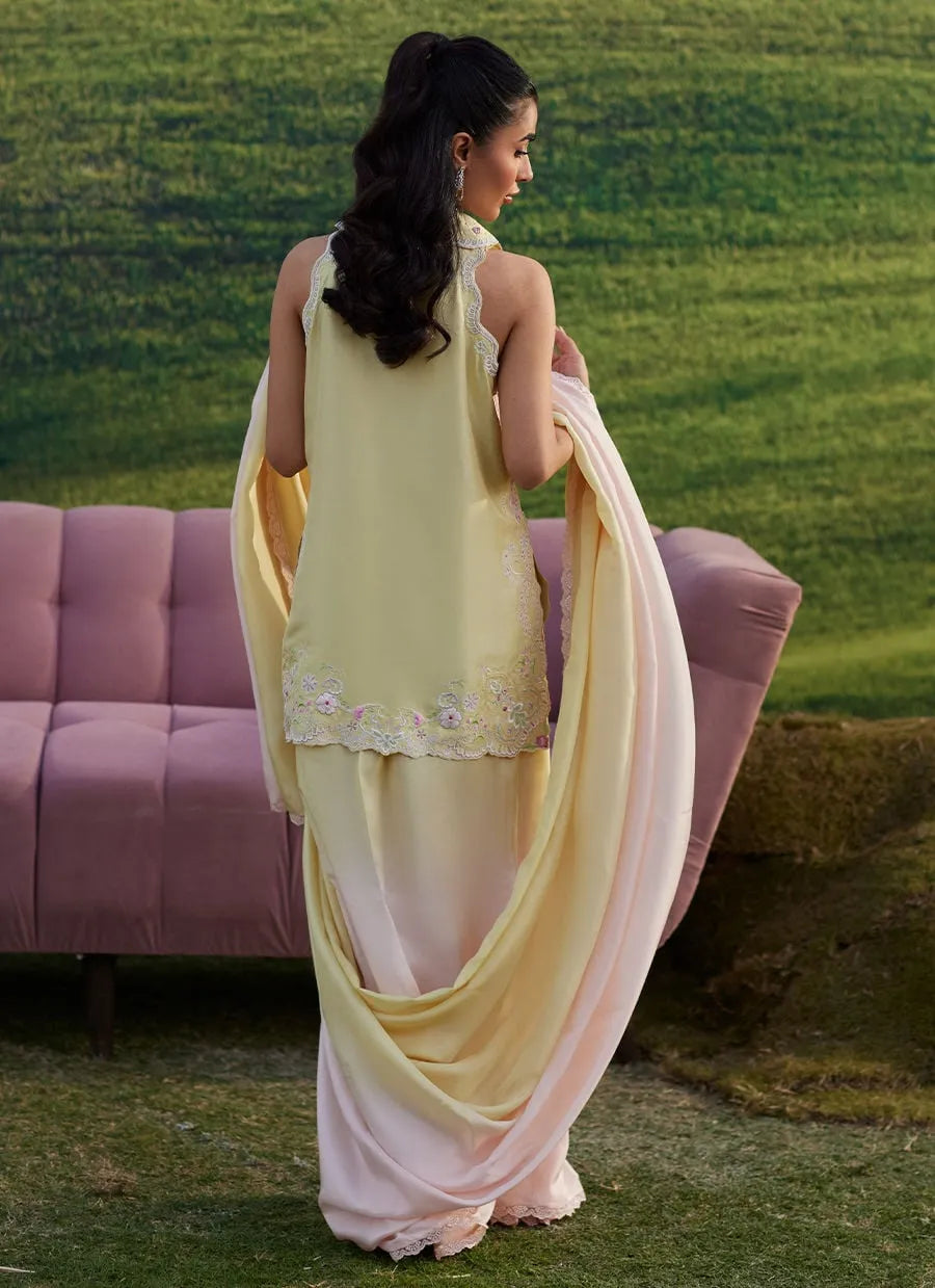 Woman in a light yellow and pink traditional outfit standing outdoors with a pink couch and green grass in the background.
