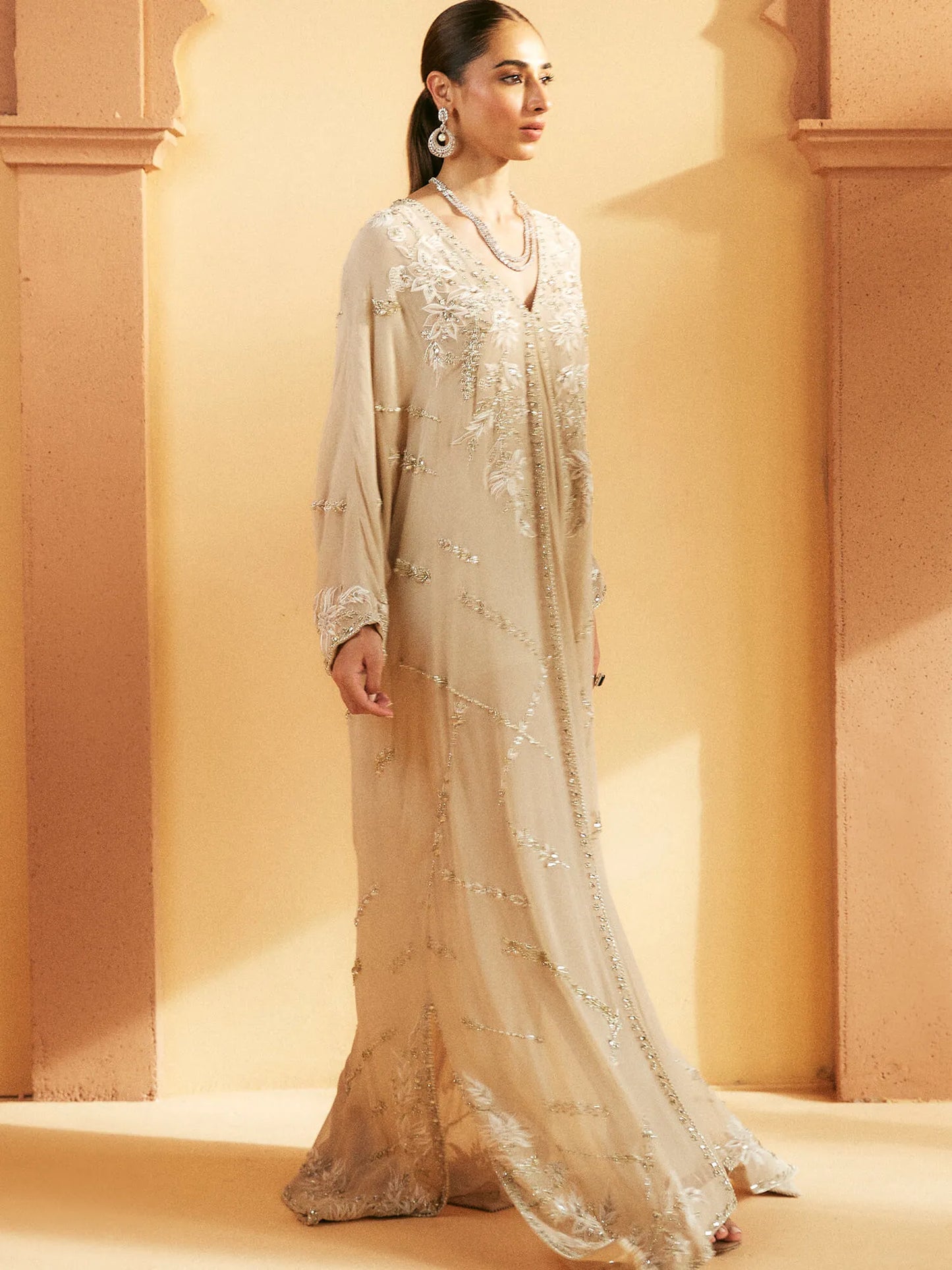 Woman wearing a light-colored embroidered dress against a warm-toned wall.
