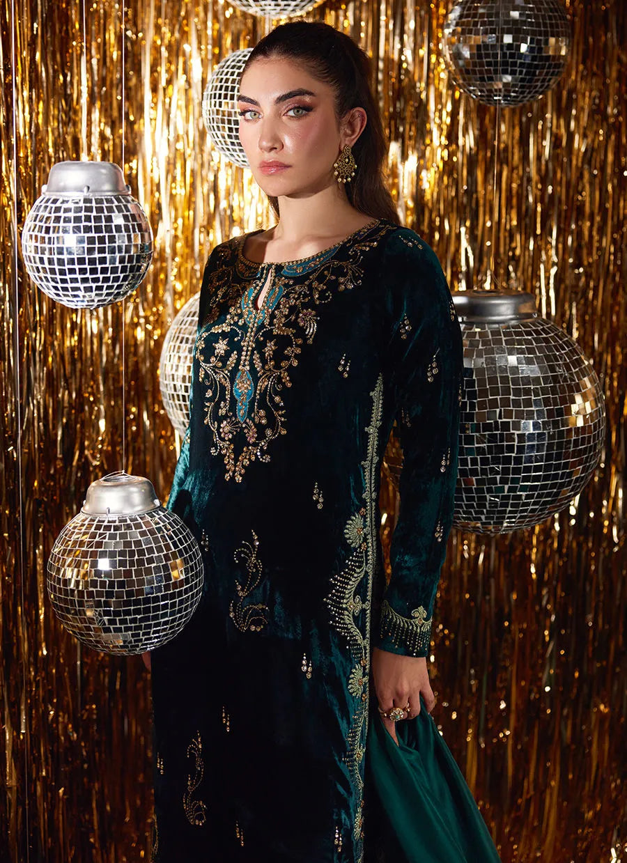Woman in a dark green embroidered dress standing against a glittery gold curtain with disco balls.