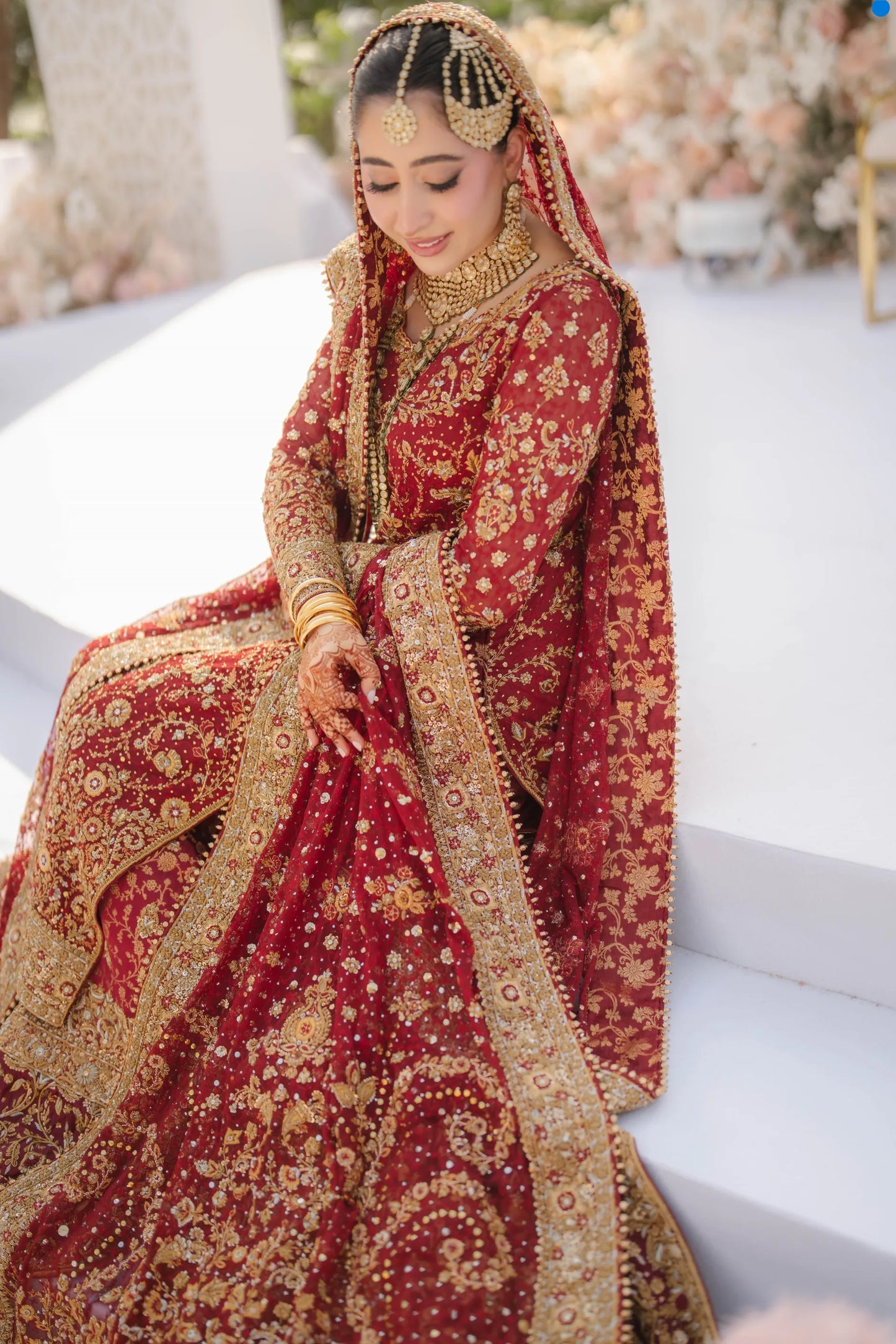 Woman in a traditional red and gold embroidered outfit with jewelry, sitting on a white surface.