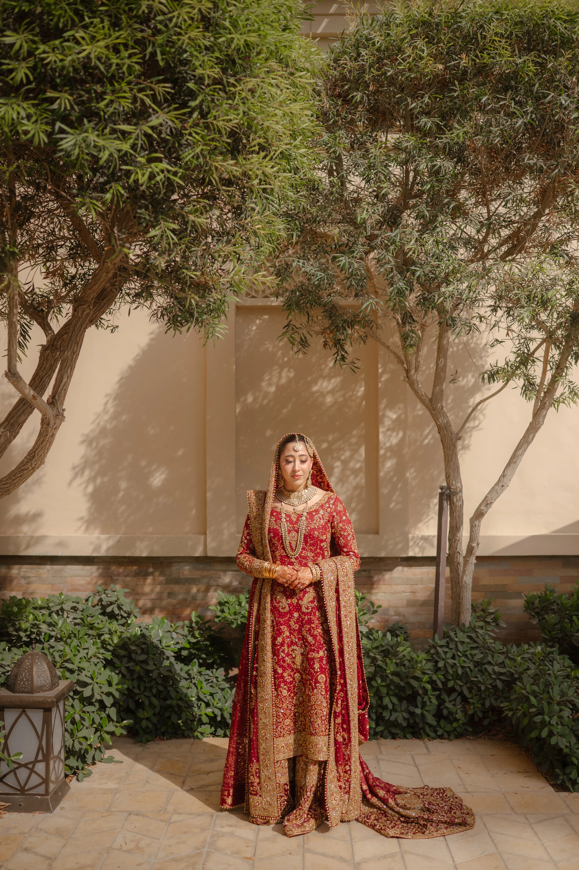 Woman in a traditional red and gold outfit standing outdoors with trees and a building in the background.