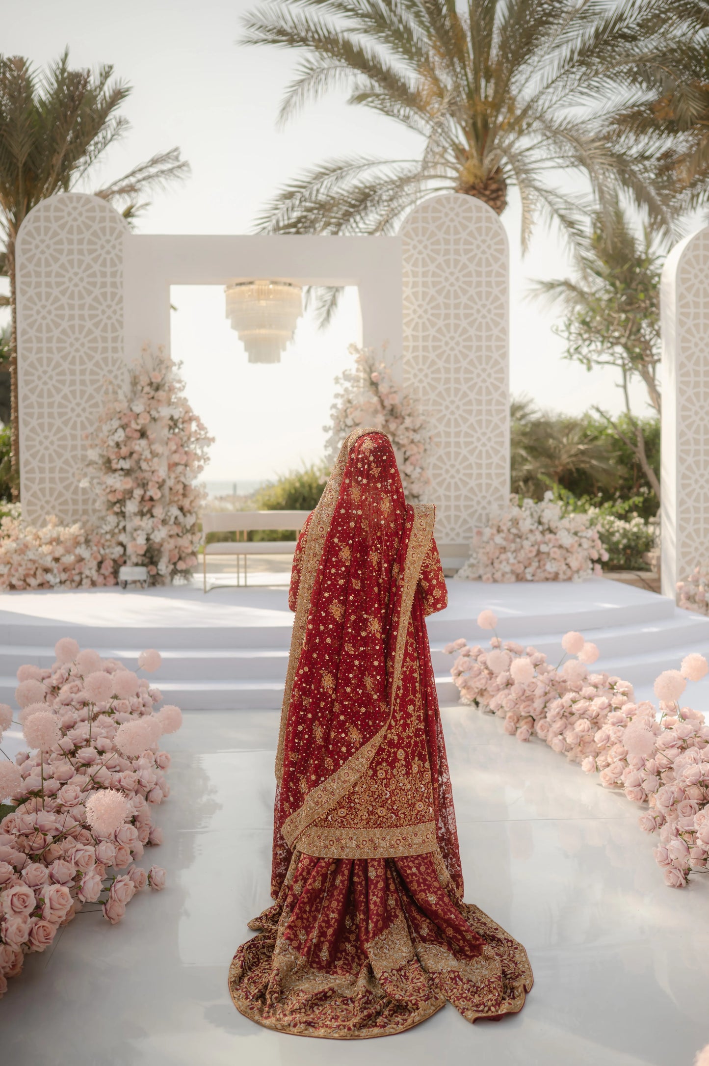 Person in a red and gold traditional outfit standing in front of a decorative archway with floral arrangements.