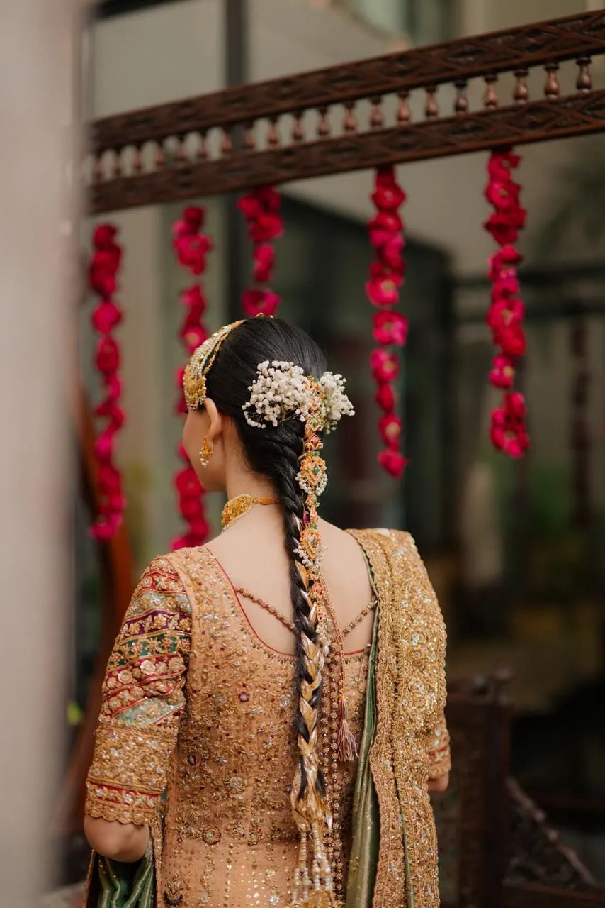 Woman in traditional attire with braided hair and floral decorations, standing in front of a decorative wall with red flowers.