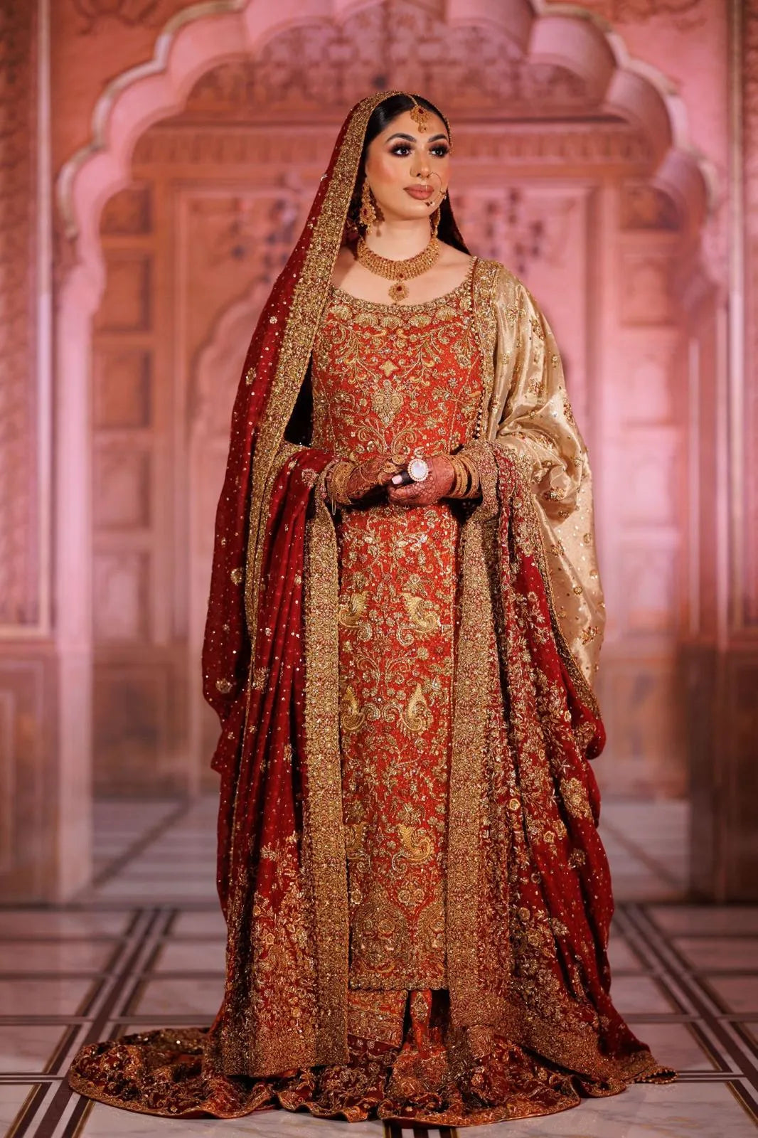 Woman in traditional red and gold bridal outfit with intricate patterns, standing against a decorative pink wall.