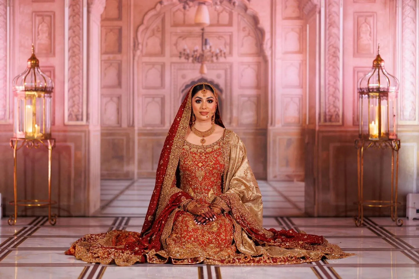 Woman in traditional red and gold outfit sitting on a patterned floor with ornate walls and lanterns.
