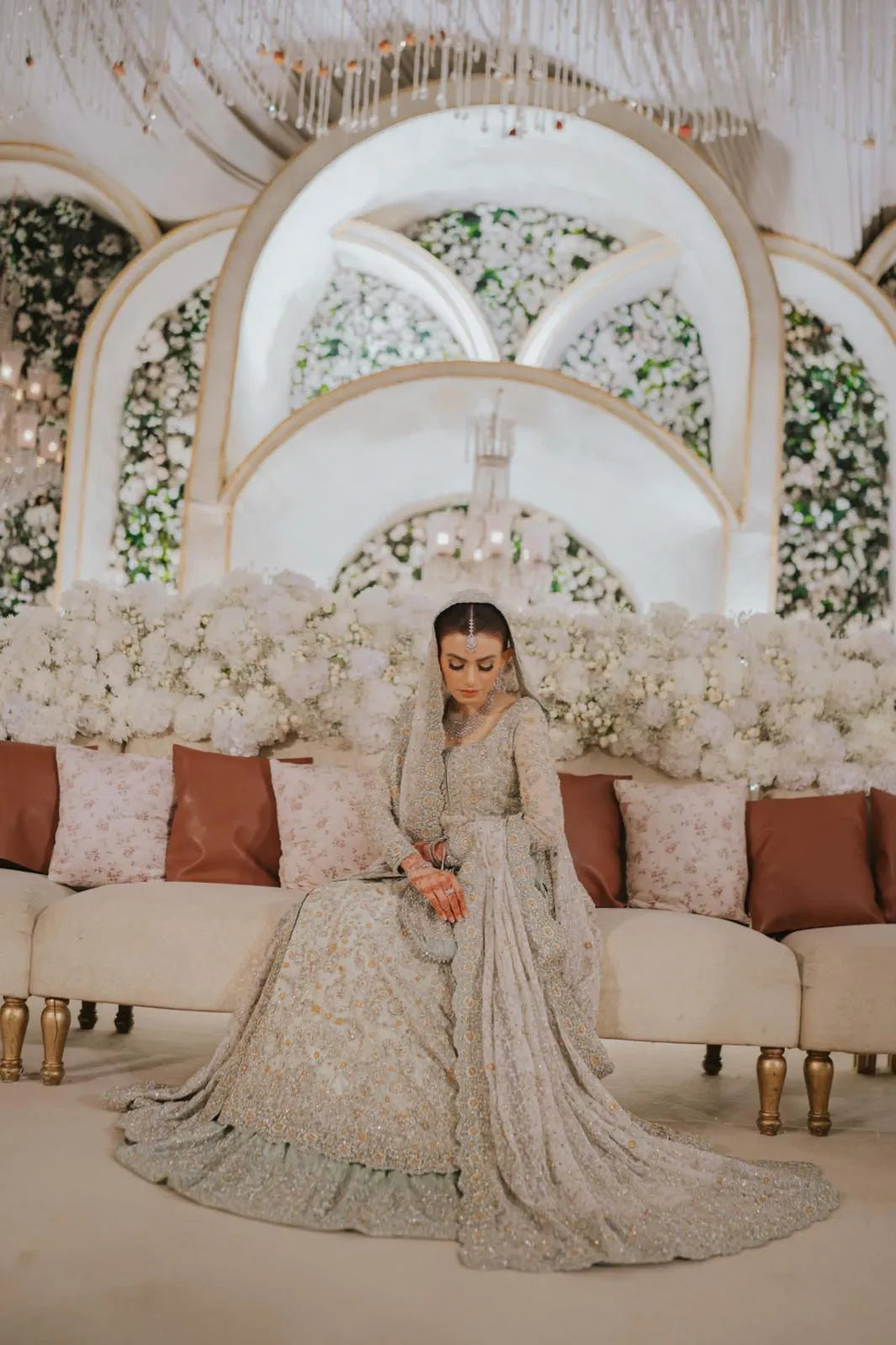 Woman in a traditional outfit sitting on a decorated stage with floral arrangements and a chandelier.