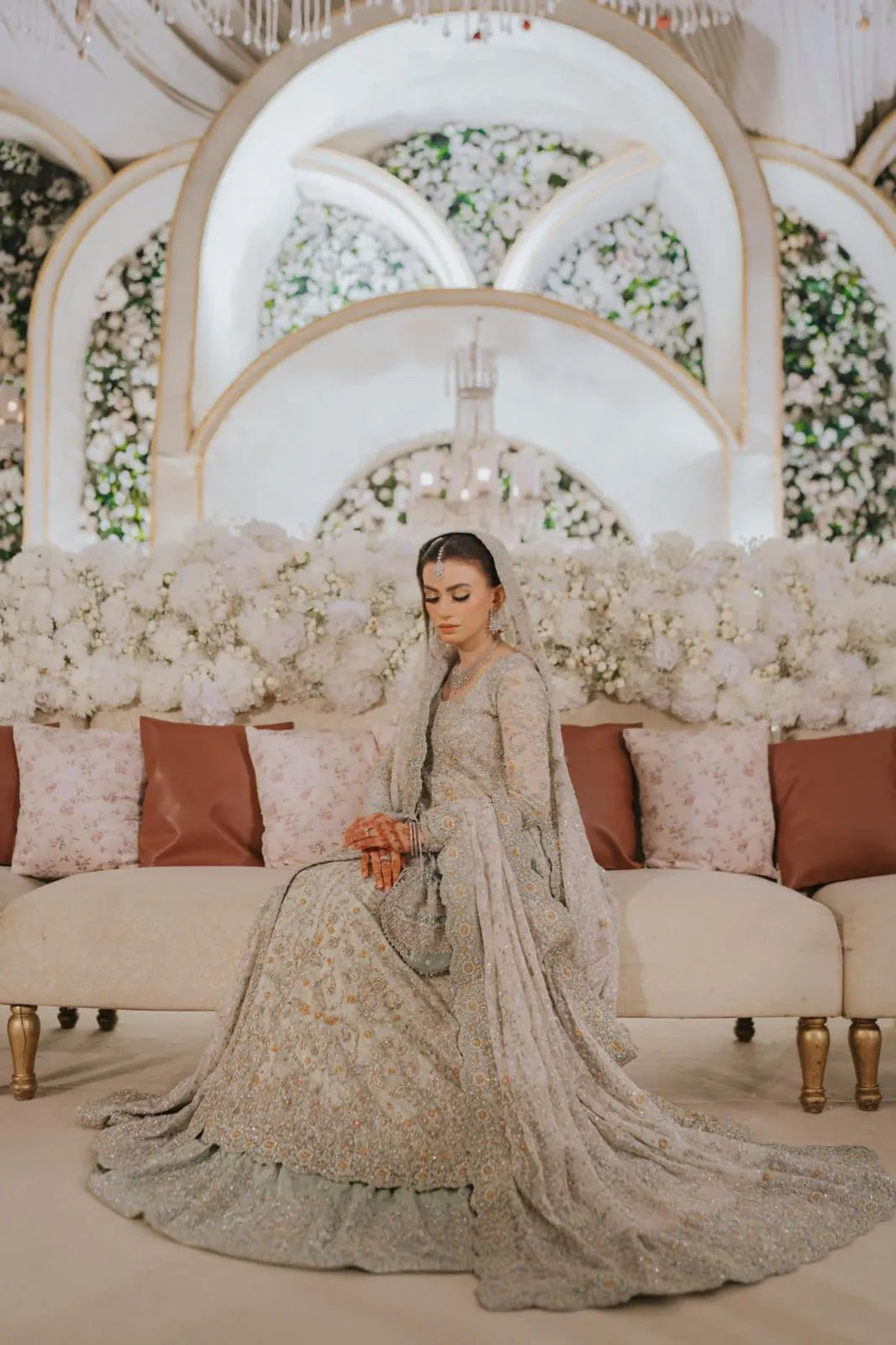 Woman in a traditional outfit sitting on a decorated sofa with floral arrangements in the background