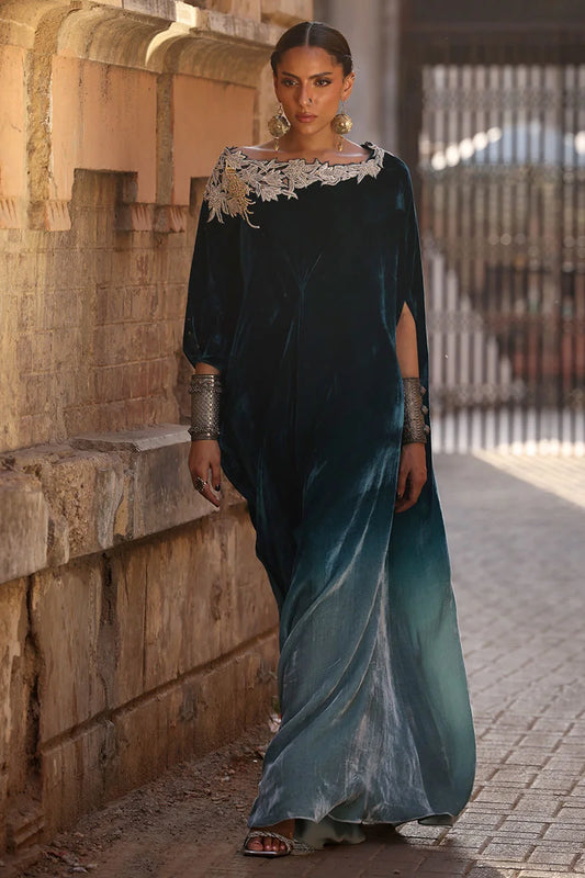 Woman in a long, dark green dress with floral details standing against a stone wall.