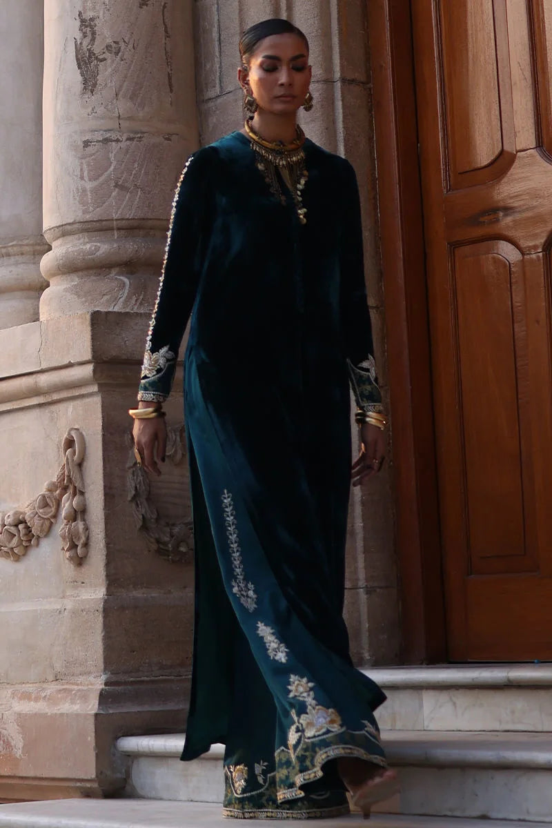 Woman in a dark green saree with floral patterns standing against a stone wall.
