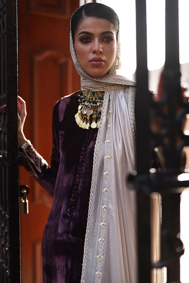 Woman in traditional attire with a white scarf and purple dress, standing in front of a black gate.