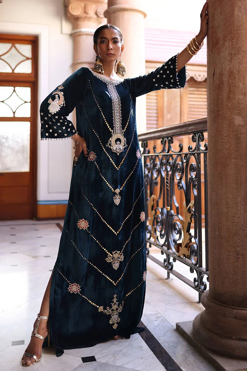 Woman in an ornate dark blue dress with gold embellishments standing in a decorative interior setting.