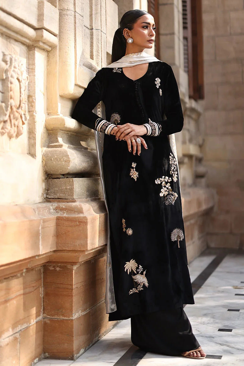 Woman in a black traditional outfit with white embroidery standing against a stone wall.