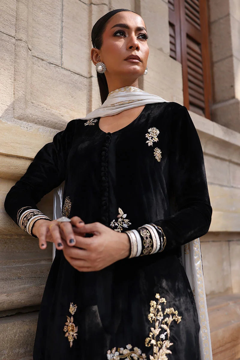 Woman in a black traditional outfit with white embroidery standing against a stone wall.