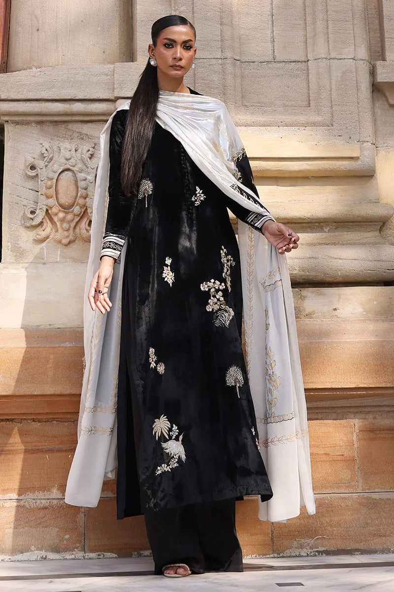 Woman wearing a black embroidered outfit with a white dupatta standing against a stone wall.
