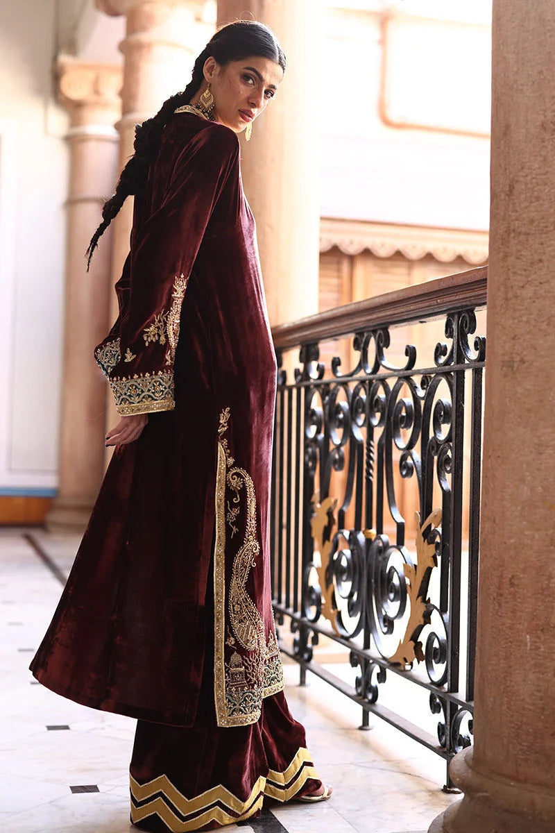 Woman in a maroon traditional outfit with gold embroidery standing in an elegant interior setting.