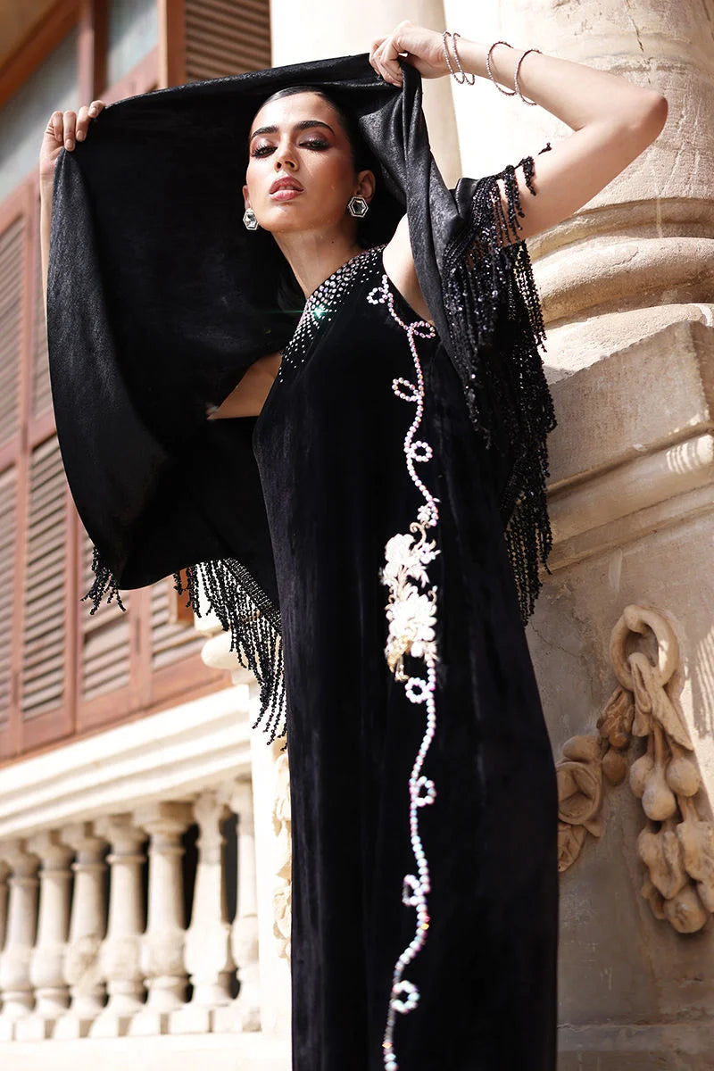 Woman in a black dress with floral embroidery standing against a stone wall.
