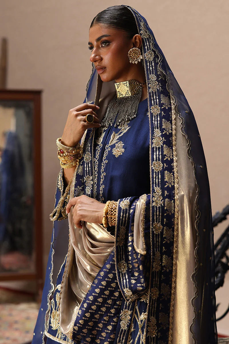 Woman in traditional blue and gold embroidered outfit with a matching dupatta, holding a pipe.