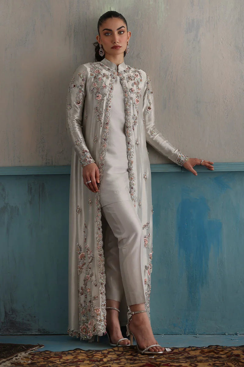 Woman wearing a long embroidered coat over a white kurta with beige pants against a textured wall.