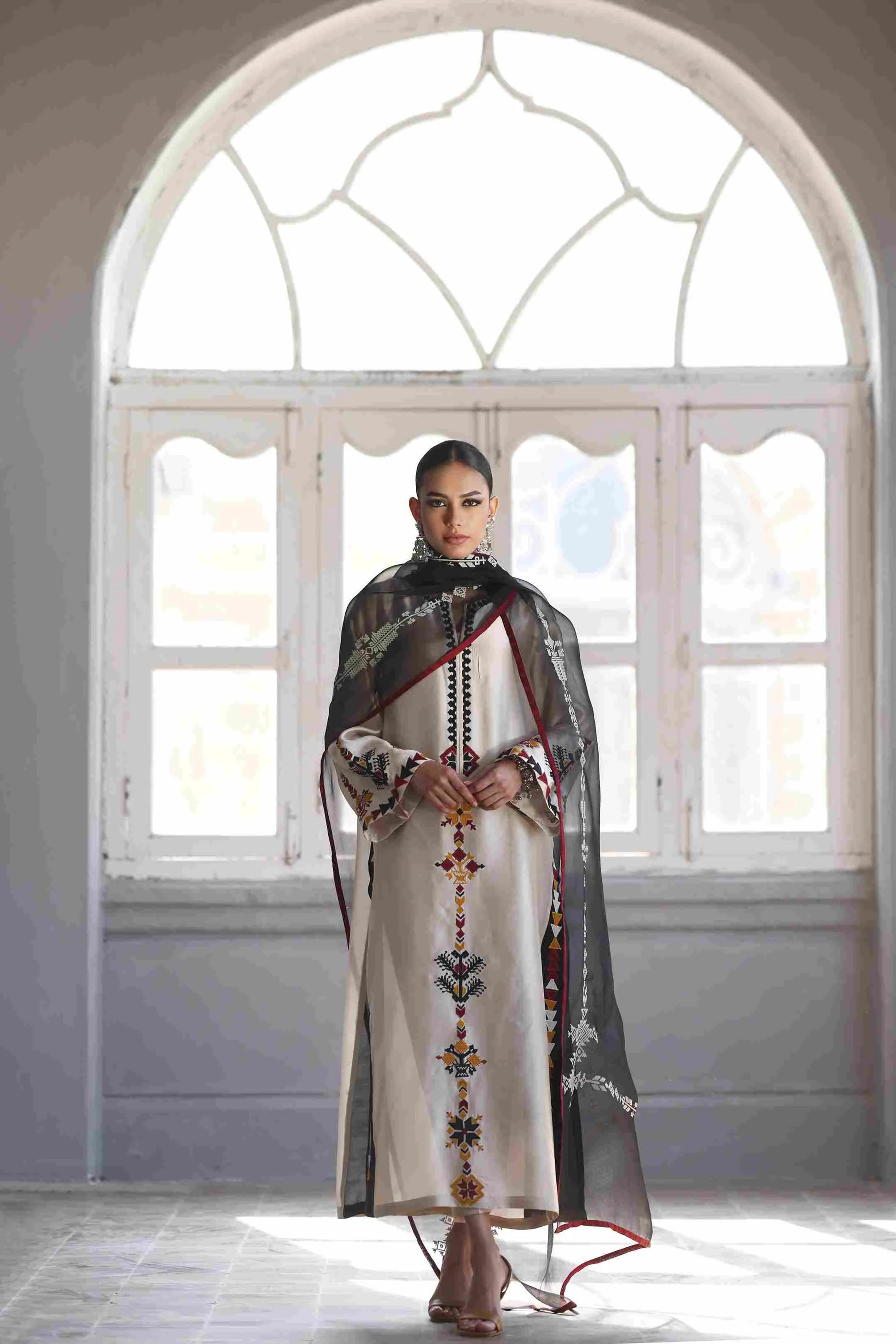 Woman in a traditional outfit standing in front of a large window with decorative glass.