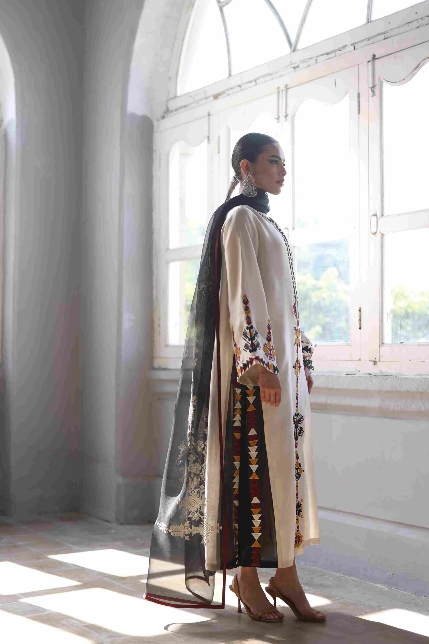 Woman wearing a long, embroidered dress standing in a bright room with large windows.