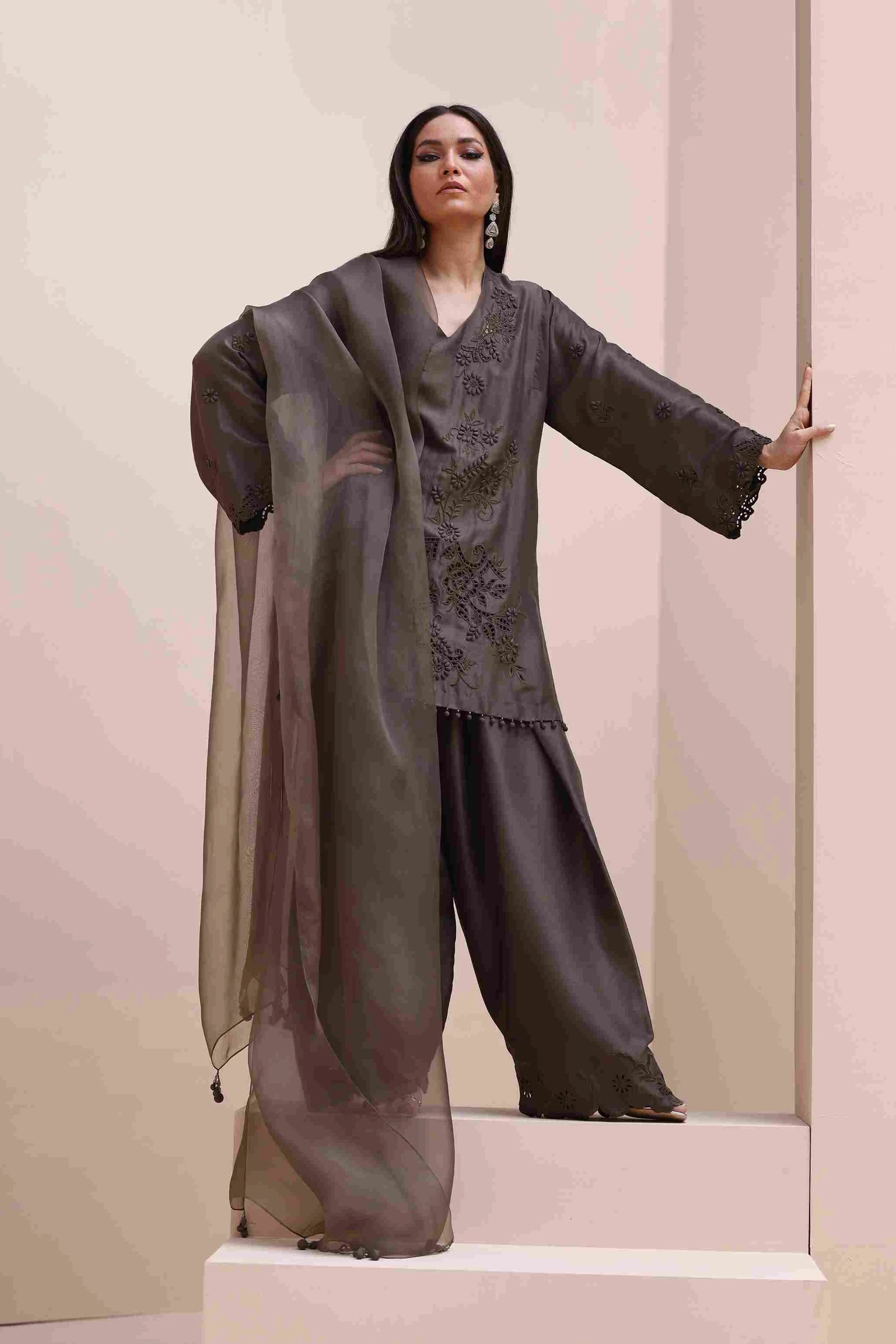 Woman wearing a dark gray traditional outfit with a sheer overlay against a light pink background